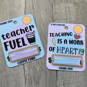 K&ouml;nnte beinhalten: Zwei Geschenkkarten mit hellblauem Hintergrund und braunem Rand. Die obere Karte zeigt eine Kaffeetasse und den Text "teacher FUEL" und "COFFEE FUND". Die untere Karte zeigt ein Herz, ein Buch und einen Stift und den Text "TEACHING IS A WORK OF HEART". Beide Karten haben einen kleinen, durchsichtigen, rechteckigen Beh&auml;lter mit einer hellblauen Fl&uuml;ssigkeit.