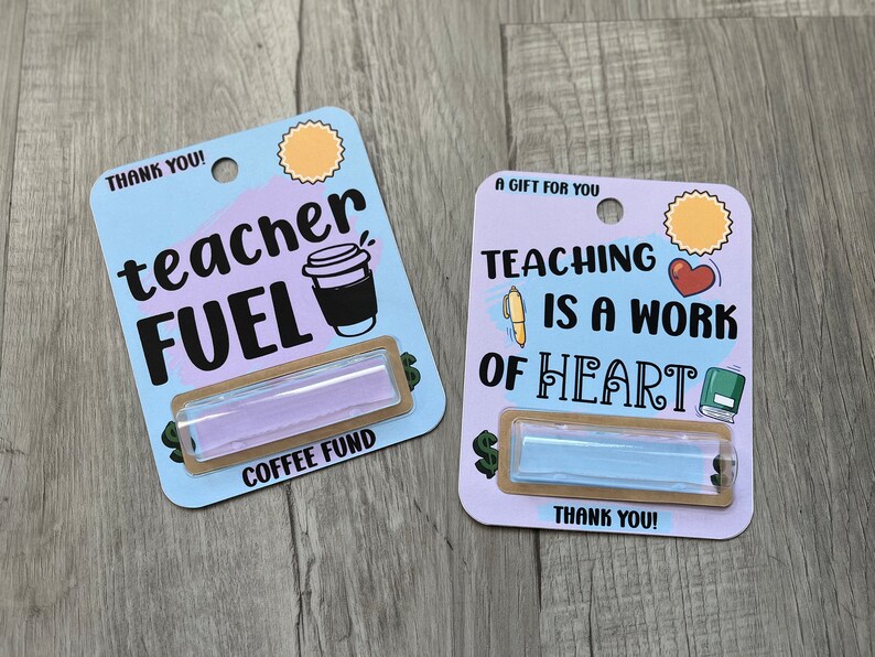 K&ouml;nnte beinhalten: Zwei Geschenkkarten mit hellblauem Hintergrund und braunem Rand. Die obere Karte zeigt eine Kaffeetasse und den Text "teacher FUEL" und "COFFEE FUND". Die untere Karte zeigt ein Herz, ein Buch und einen Stift und den Text "TEACHING IS A WORK OF HEART". Beide Karten haben einen kleinen, durchsichtigen, rechteckigen Beh&auml;lter mit einer hellblauen Fl&uuml;ssigkeit.