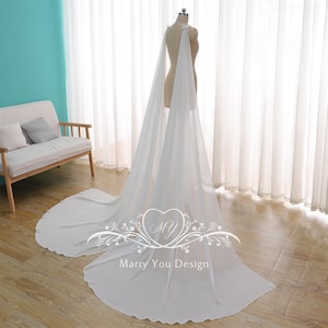 May include: A white sheer bridal cape with a scalloped edge. The cape is draped over a dress form and is flowing on the floor. The text "Marry You Design" is printed on the cape.