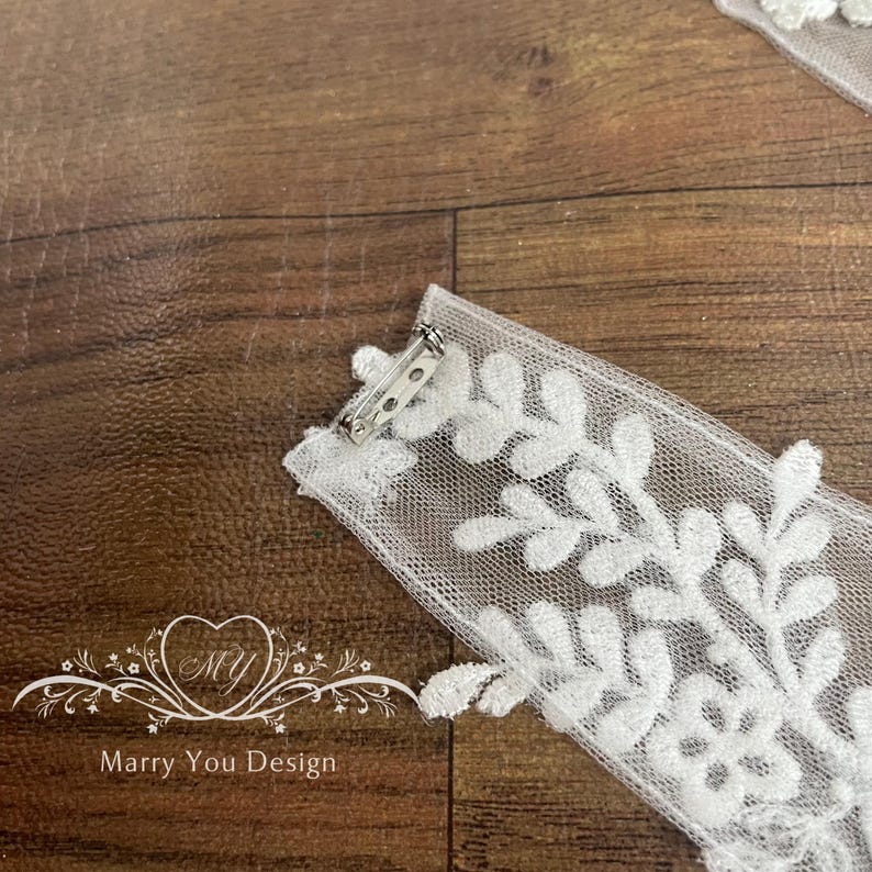 Puede incluir: Primer plano de una aplicaci&oacute;n de encaje blanco con dise&ntilde;o floral y un alfiler plateado. La aplicaci&oacute;n est&aacute; sobre una superficie de madera. El texto "Marry You Design" es visible.