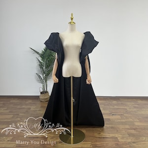 Puede incluir: Una capa negra con grandes mangas abullonadas y dramáticas. La capa se muestra en un maniquí con un soporte dorado. La capa es de largo hasta el suelo y tiene una silueta fluida.