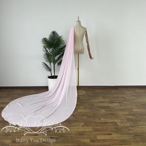 May include: A long, flowing pink tulle veil with a scalloped edge. The veil is draped over a dress form and is displayed on a wooden floor.