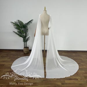 May include: A white chiffon bridal cape with a long train. The cape has a simple design with a gathered neckline and a flowing silhouette.