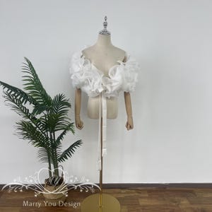 Puede incluir: Bolero de novia blanco de hombros descubiertos con detalles de volantes y cierre de lazo. El bolero se muestra en un maniquí blanco.