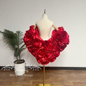 May include: A vibrant red, rose-adorned garment displayed on a mannequin. The garment is crafted from a satin-like material, with numerous fabric roses creating a voluminous, heart-shaped design. The mannequin is on a gold stand, with a plant in a white pot in the background.