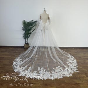 May include: A long, white bridal veil with a floral lace border. The veil is displayed on a mannequin, showcasing its flowing design. The bottom edge features intricate white floral embroidery, adding a delicate touch to the wedding accessory.