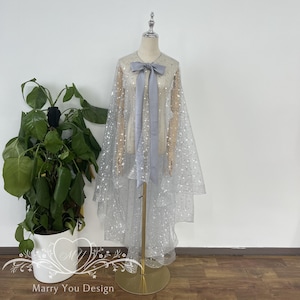 May include: A silver tulle cape with a silver glitter pattern and a light gray satin bow. The cape is on a mannequin and has a high-low hemline.