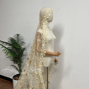 May include: A cream-colored bridal veil featuring intricate gold star and celestial embroidery, accented with small sequins. The veil has a scalloped lace trim with a star-shaped pattern along the edges and is displayed on a mannequin.