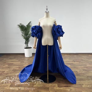 May include: A royal blue dress with a long train and large, puffy sleeves. The dress is on a mannequin and is displayed against a white wall.