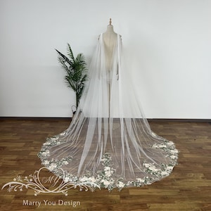 May include: A white tulle wedding veil with a floral design and pearl embellishments. The veil is long and flowing, with a cape-like back.