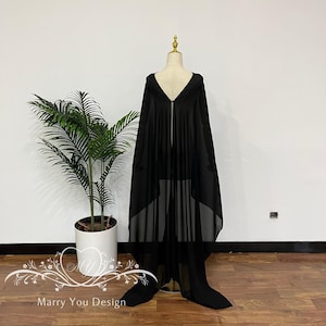 Op de afbeelding: Een zwarte cape met capuchon met een lang, vloeiend ontwerp. De cape wordt getoond op een mannequin, die de draperie en textuur laat zien. De stof lijkt licht en semi-transparant, met een ritssluiting aan de voorkant.