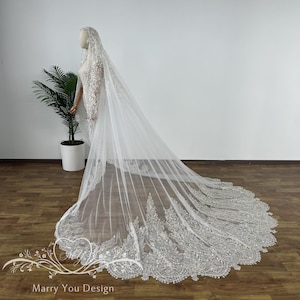 May include: A long, white bridal veil with intricate lace detailing along the edges. The veil is draped over a mannequin, showcasing its length and design. The lace features a detailed floral pattern.