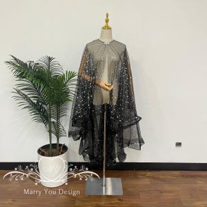 May include: A black cape with silver star-shaped embellishments, displayed on a mannequin. The cape features a gradient effect, transitioning from a lighter top to a darker bottom. A decorative clasp secures the cape at the neck.