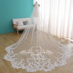 May include: A long, white bridal veil with a delicate floral lace trim. The veil is displayed on a mannequin, showcasing its length and intricate design. The veil's edge features a detailed leaf pattern.