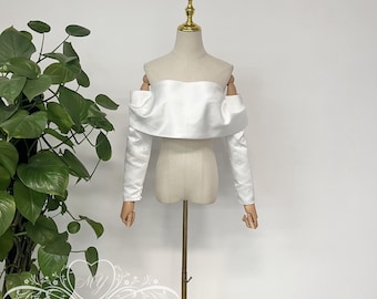 May include: White off-the-shoulder bridal bolero with long sleeves and a ruffled neckline. The bolero is displayed on a white mannequin.