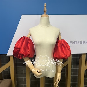 May include: A pair of red satin puff sleeves on a white dress form. The sleeves are gathered at the top and have a loose, flowing fit.