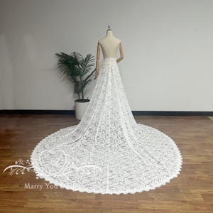 Op de afbeelding: Een witte kanten bruidsjurk-sleep die op een mannequin wordt getoond. De sleep is lang en cirkelvormig, met een geschulpte rand. De jurk wordt getoond voor een witte muur en een plant in een pot. De tekst "Marry You" is zichtbaar.