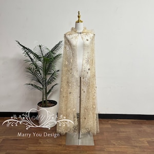 May include: A sheer, beige cape with a star and snowflake pattern, displayed on a mannequin. The cape features a high collar and is embellished with sequins. The cape is floor-length and has a flowing design, suitable for special occasions.