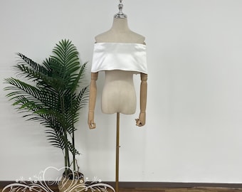 May include: A white off-the-shoulder satin bridal bolero on a dress form. The bolero is on a gold stand against a white wall.