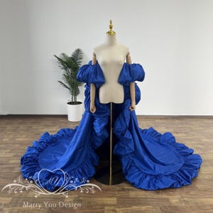 May include: A royal blue gown with a dramatic, ruffled train and off-the-shoulder sleeves is displayed on a mannequin. The gown features a voluminous design and is set against a neutral background with a potted plant.