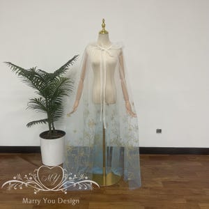 May include: A sheer, white cape with a hood and gold floral embroidery. The cape is displayed on a mannequin with a gold stand. The bottom of the cape has a gold floral design. The words "Marry You Design" are at the bottom.