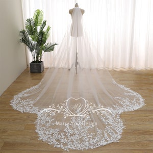 May include: A long, white bridal veil with intricate floral lace detailing along the edges. The veil is displayed on a mannequin, showcasing its flowing design. The delicate lace adds a touch of elegance to the bridal accessory.
