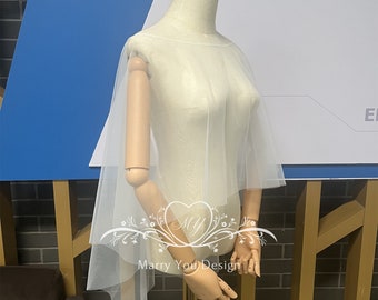Puede incluir: Una capa de novia de tul blanco con un borde festoneado. La capa está drapeada sobre un maniquí y tiene un diseño simple y elegante.
