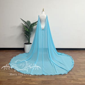 May include: A light blue, flowing cape with a long train, displayed on a mannequin. The cape has a deep V-back and is made of a sheer fabric. The train spreads out on the floor. The cape is designed for special occasions.