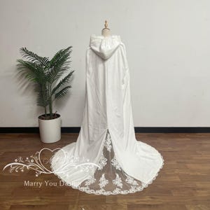 May include: A white hooded cloak with a long train, displayed on a mannequin. The cloak features a lace-trimmed hem and a sheer panel at the back. The hood is adorned with delicate lace detailing. The cloak is made of a soft, flowing fabric.
