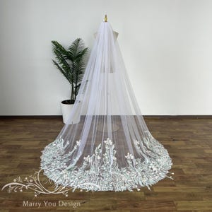 Peut inclure: Un long voile de mariée blanc avec un motif floral sur le bord inférieur. Le voile est fait d'un tissu transparent et est présenté sur un mannequin. Le motif floral présente des accents blancs et bleu clair. Le texte "Marry You Design" est visible.