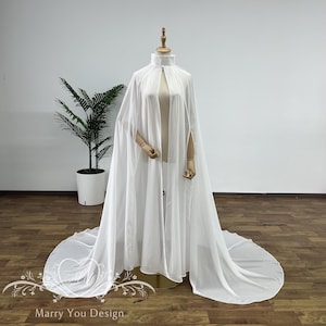 Op de afbeelding: Een witte, vloeiende cape met een hoge kraag en open voorkant, gepresenteerd op een mannequin. De cape heeft lange, elegante mouwen en een sleep die zich over de vloer uitstrekt. De tekst "Marry You Design" is onderaan zichtbaar.