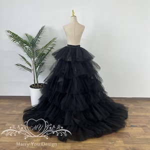 Puede incluir: Falda de tul negra con múltiples capas, exhibida en un maniquí. La falda tiene una larga cola y una cintura ancha. El maniquí está sobre un suelo de madera, con una planta en maceta a la izquierda.