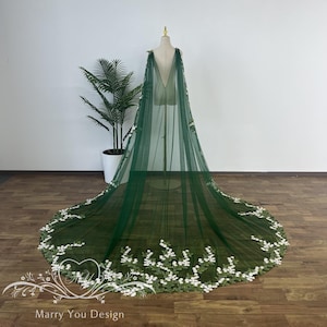May include: A dark green tulle cape with a long train, displayed on a mannequin. The cape is adorned with white floral embroidery along the hemline and shoulder area. The cape is a wedding or formal wear item.
