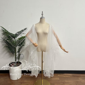 Può includere: Una mantellina da sposa in tulle bianco trasparente con un delicato bordo arricciato. Il tessuto leggero e fluido si drappeggia elegantemente sulle spalle di un manichino, creando un aspetto morbido e stratificato con una finitura traslucida.
