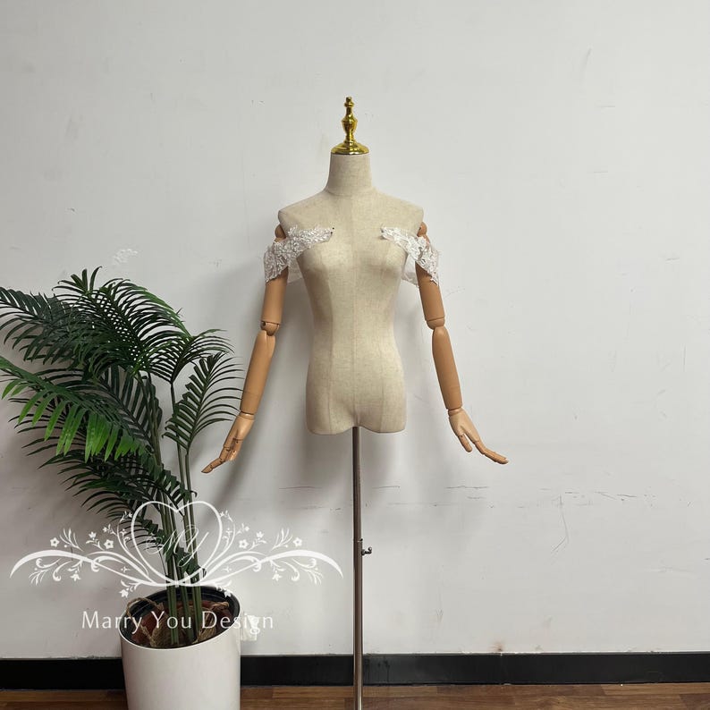 Puede incluir: Un maniqu&iacute; beige con brazos articulados de madera y cuello dorado. Delicada encaje blanco adorna los hombros. El maniqu&iacute; est&aacute; montado sobre un soporte met&aacute;lico. El texto "Marry You Design" es visible en la esquina inferior izquierda.