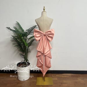 May include: A large, dusty rose-colored satin bow with long, cascading fabric tails attached to a beige mannequin torso. The bow features a smooth, reflective finish and is mounted on a gold-toned metal stand with a square base.