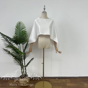 May include: A white satin bridal cape with a short, rounded neckline and a flowing silhouette. The cape is displayed on a mannequin.