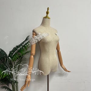 Puede incluir: Un maniqu&iacute; beige con brazos de madera articulados y una parte superior dorada. Un detalle de manga y hombro de encaje blanco est&aacute; prendido al maniqu&iacute;. El texto "Marry You Design" es visible en la esquina inferior izquierda.