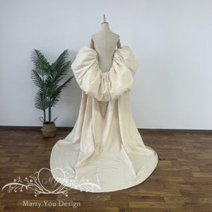 Peut inclure: Une robe de mariée crème avec un détail de dos spectaculaire et surdimensionné. La robe présente un design volumineux et froncé aux épaules et dans le haut du dos, se transformant en une longue et élégante traîne. La robe est présentée sur un mannequin en studio.
