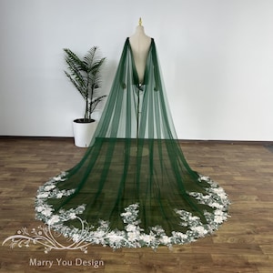 May include: A dark green tulle cape with a floral lace trim. The cape is displayed on a mannequin, with the train extending across the floor. The floral trim is white and adds a delicate contrast to the dark green fabric. The cape is designed for special occasions.
