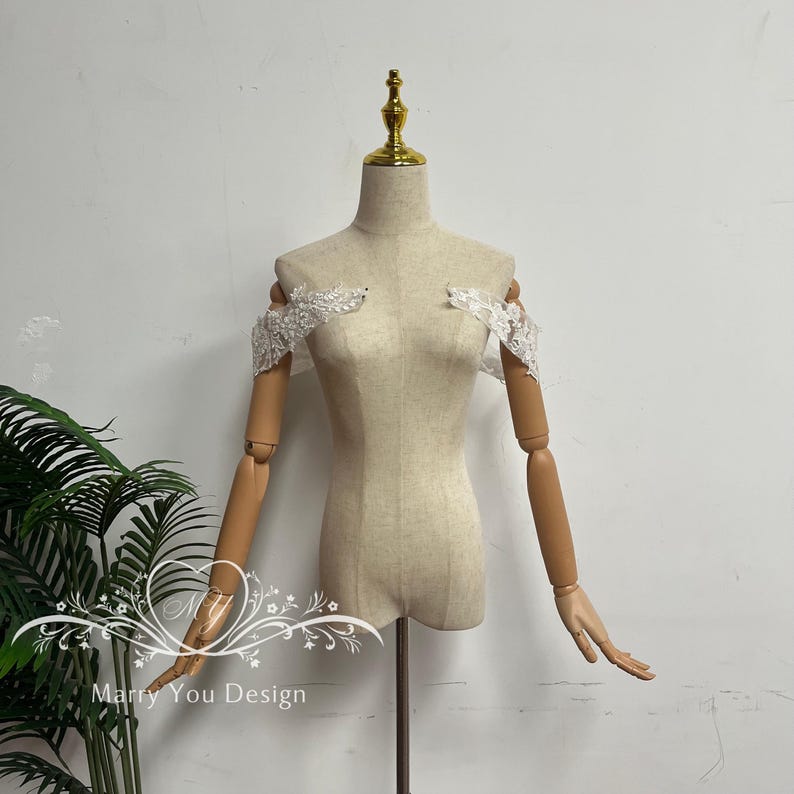 Puede incluir: Un maniqu&iacute; beige con brazos articulados de madera y una parte superior dorada. Delicadas piezas de encaje blanco para los hombros est&aacute;n sujetas al maniqu&iacute;. El texto "Marry You Design" es visible en la esquina inferior izquierda.
