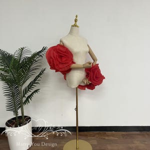 Puede incluir: Un maniquí con torso beige y brazos de madera, adornado con grandes adornos de tela roja en forma de rosa en los hombros y la cintura. El maniquí está sobre un soporte de metal dorado. Hay una planta en maceta en el fondo. El texto "Marry You Design" es visible.