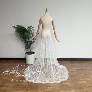 May include: A white lace skirt with a long train is displayed on a mannequin. The skirt is sheer, revealing the mannequin's form. The lace features a delicate floral pattern. The mannequin is positioned in front of a white wall and a potted plant.