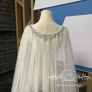 Puede incluir: Una capa de novia de tul blanco con un escote de cuentas. Las cuentas son plateadas y tienen un diseño de cadena colgante. La capa es transparente y tiene una silueta fluida. El texto "Marry You Design" es visible en la parte inferior de la imagen.