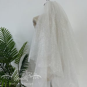 May include: A white tulle wedding veil with a sparkly finish. The veil is draped over a mannequin and has a soft, flowing texture.