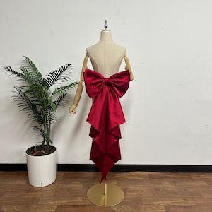 May include: A deep red satin fabric accessory featuring a large, structured bow at the top with long, cascading, pleated tails that drape downward. The accessory is displayed on a beige mannequin torso with articulated wooden arms and a gold base.