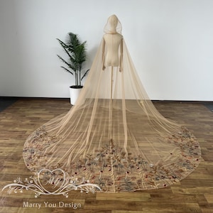 May include: A beige hooded cape with an embroidered floral design. The cape is displayed on a mannequin and has a long train. The floral embroidery features red, blue, and green accents. The text "Marry You Design" is visible at the bottom.