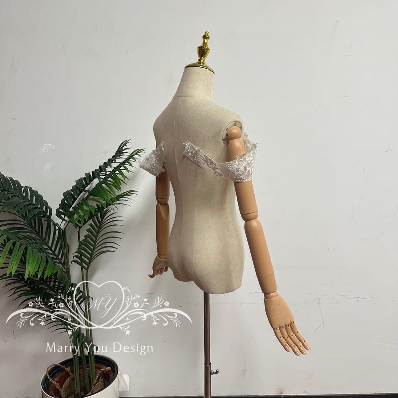 Puede incluir: Un maniqu&iacute; beige con brazos articulados de madera y una pieza para la cabeza dorada. El maniqu&iacute; est&aacute; adornado con un detalle de hombro de encaje blanco. Una planta en maceta est&aacute; en la esquina inferior izquierda. El texto "Marry You Design" es visible.