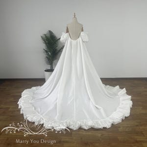 Könnte beinhalten: Ein weißes Brautkleid mit langer Schleppe und Rüschensaum wird auf einer Schaufensterpuppe präsentiert. Das Kleid hat einen offenen Rücken, schulterfreie Ärmel und einen fließenden Rock. Das Kleid ist aus einem glänzenden, satinartigen Stoff gefertigt.