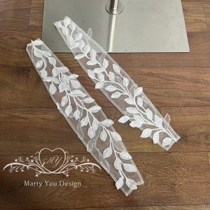 May include: Two white, leaf-patterned lace appliques. The delicate, sheer fabric is adorned with detailed, raised leaf designs. The appliques are displayed on a wooden surface. The text "Marry You Design" is visible.